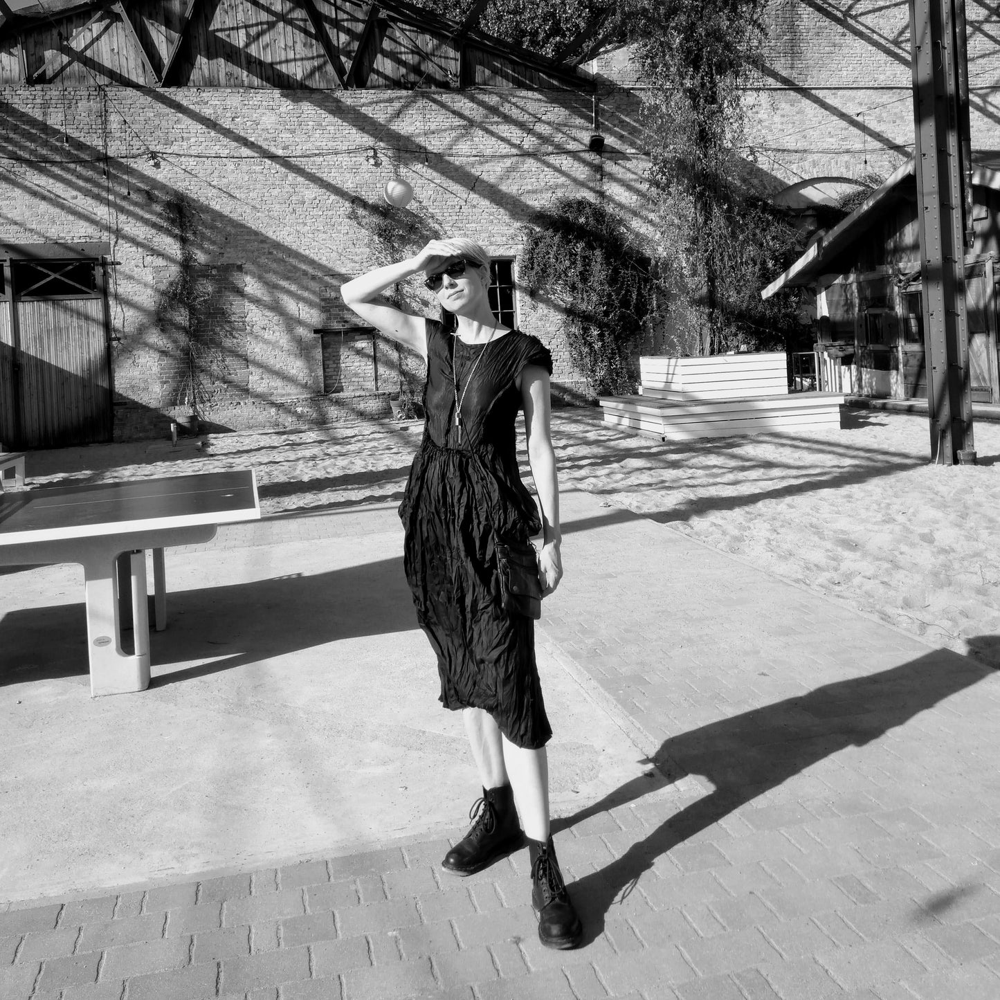 A figure stands in a minimalist outdoor space, wearing a black, textured dress and combat boots, with shadows creating a dynamic backdrop.