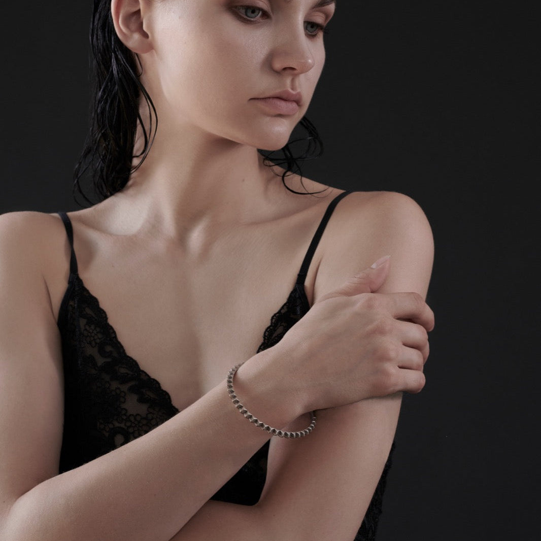 A woman with wet, slicked-back hair wears a black lace top and silver bracelet, gently embracing her arm against a dark backdrop.
