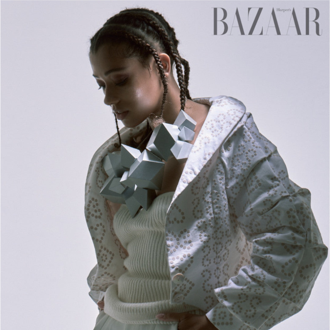 A model wearing a stylish white jacket with intricate patterns and a bold geometric necklace, captured in an artistic pose.