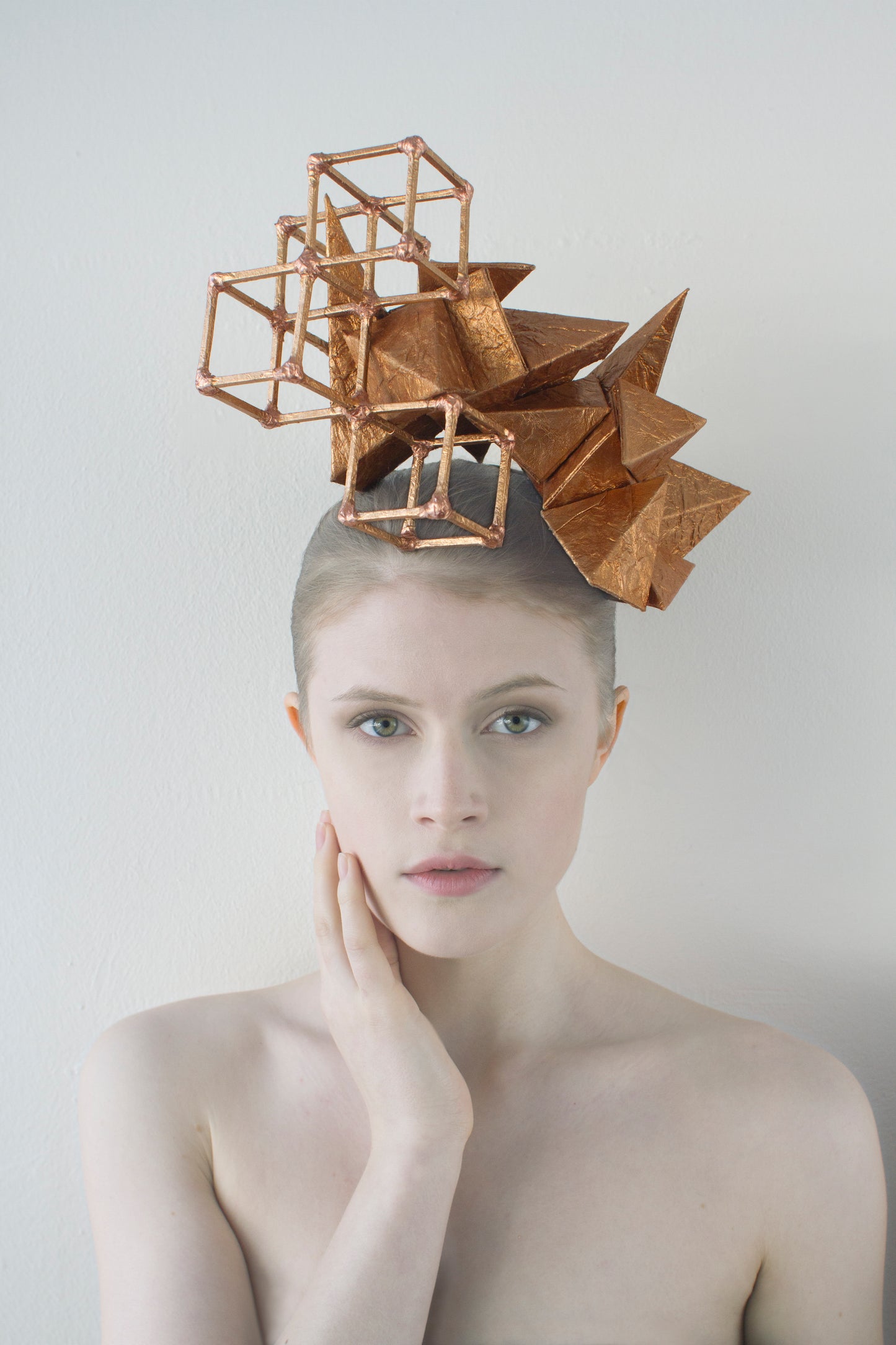 A woman sitting in front of white background touching her face with her right hand wears a copper-colored geometric headpiece made from pyramids and cubes.