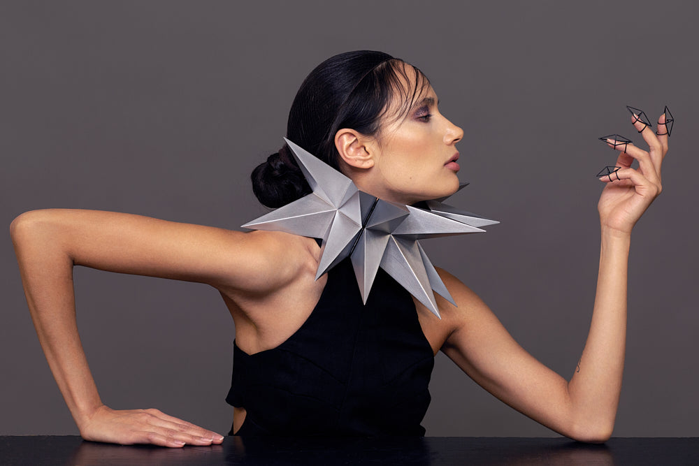 A model wearing a striking, origami-inspired silver collar and dramatic elongated black nails poses against a neutral background.