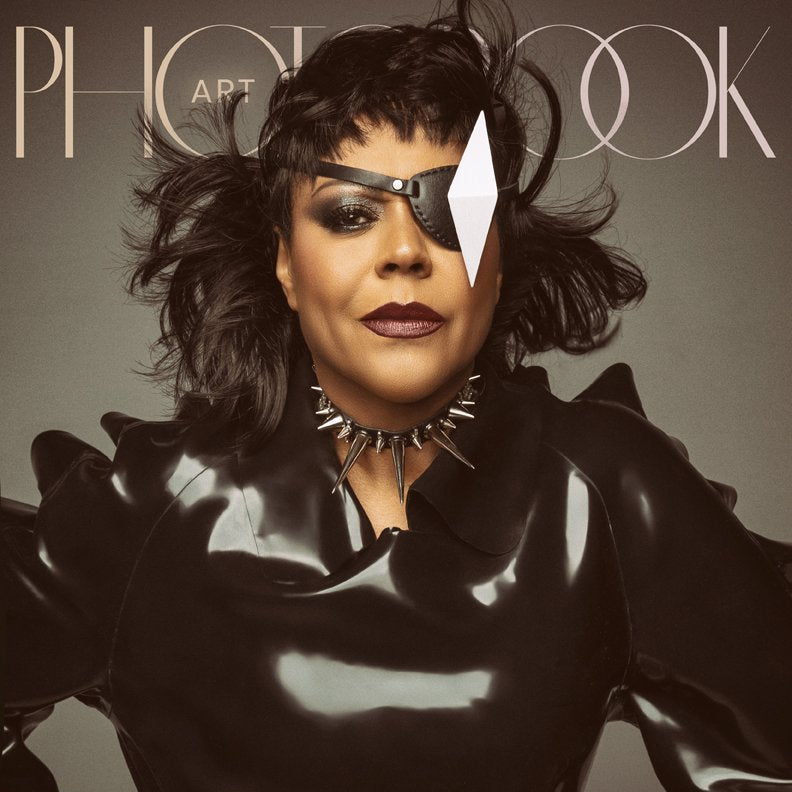 A stylish individual wears a glossy black outfit with bold shoulder details, a geometric eyepatch, and a striking spiked necklace, on the cover of PhotoBook Magazine.