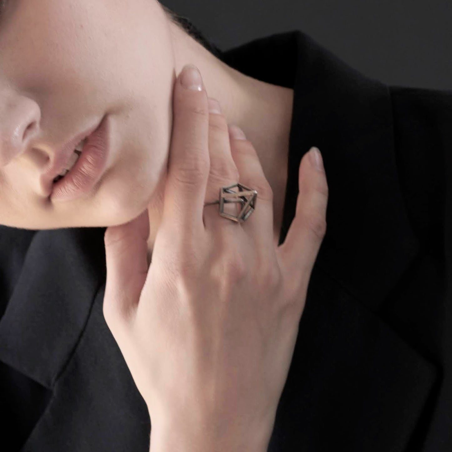 Close-up of a woman's hand touching her chin, wearing a large, geometric ring, with a blurred black jacket background.