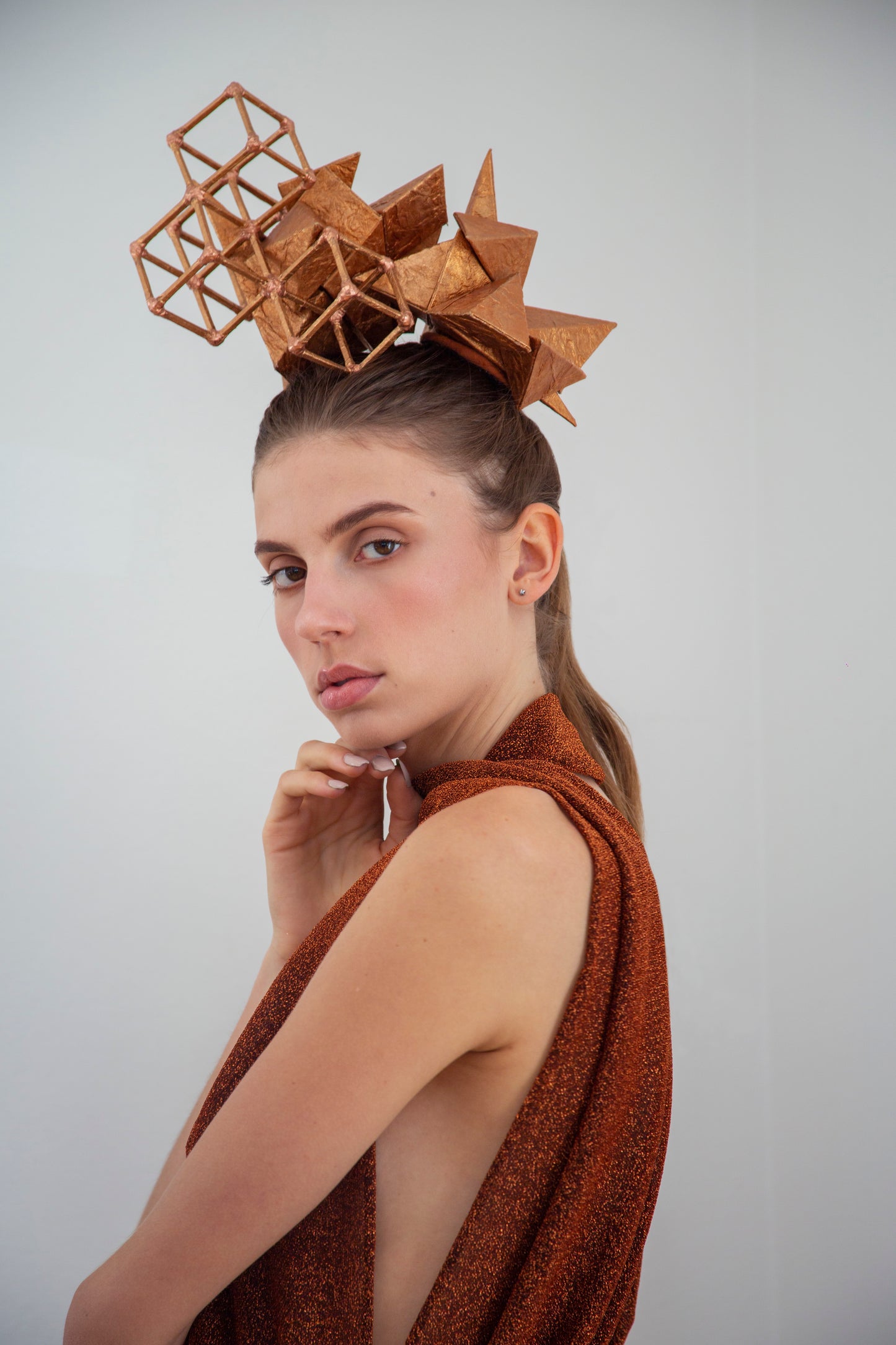 A woman in a copper-colored shirt looks to her left, touching her chin with her right hand and wears a copper-colored geometric headpiece made from pyramids and cubes.