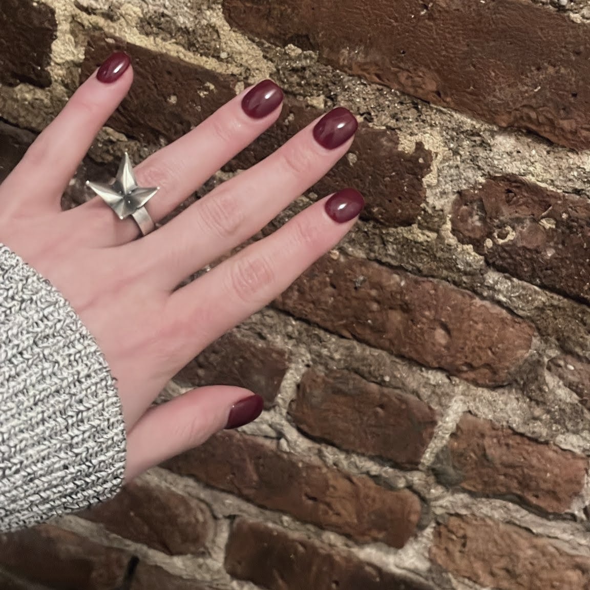 A hand with dark red nails and a silver star-shaped ring rests against a rustic brick wall.