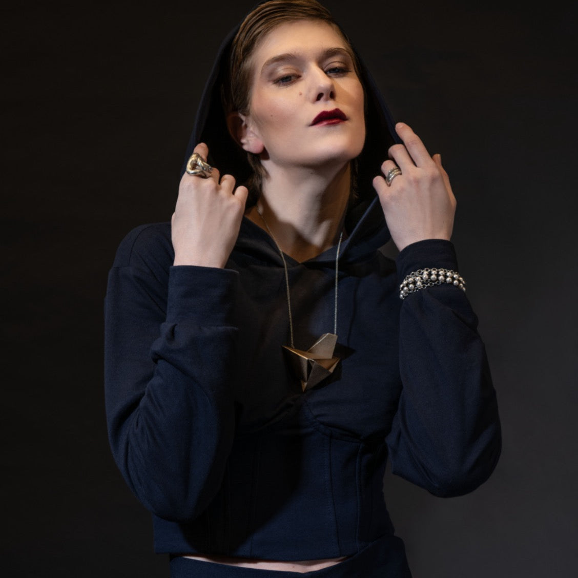 A model in a navy crop hoodie poses confidently against a dark background, showcasing bold rings and a sculptural necklace.