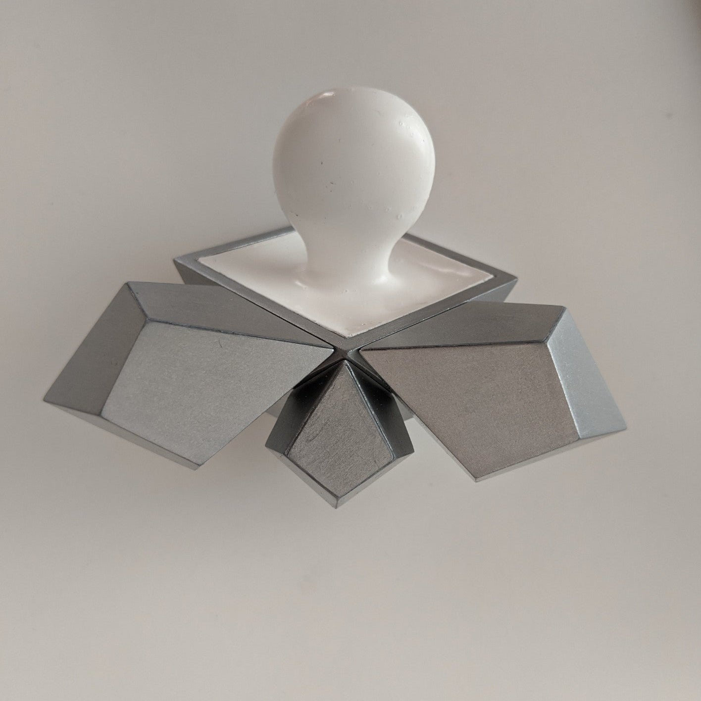 A white spherical object atop a silver geometric base featuring multiple angular shapes, set against a plain background.