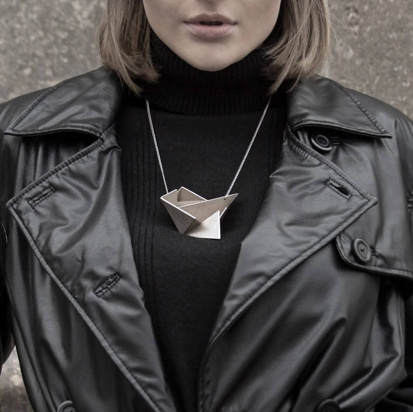 A person wearing a black leather jacket and a black turtleneck, showcasing a unique geometric silver necklace against a textured background.