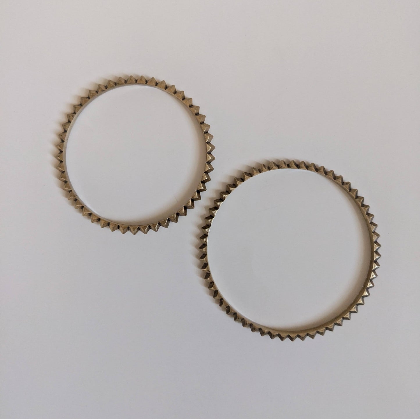 Two circular brass bracelets with a zigzag edge design, set against a light grey background.