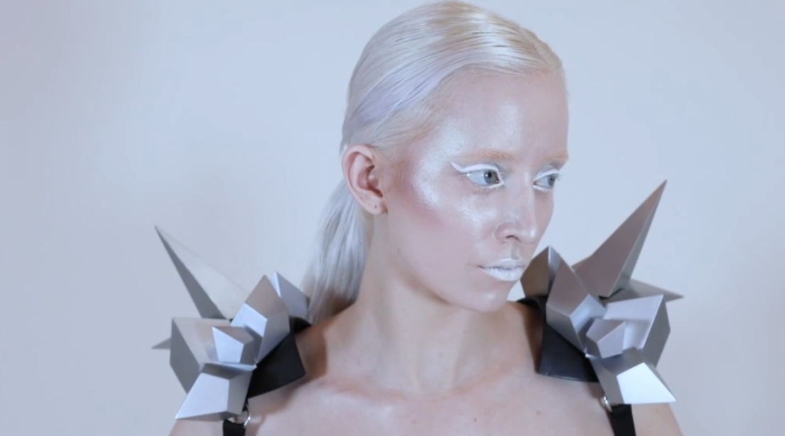 A pale woman wearing white gracefully moves around while wearing a variety of silver geometric avant garde jewelry and accessories.