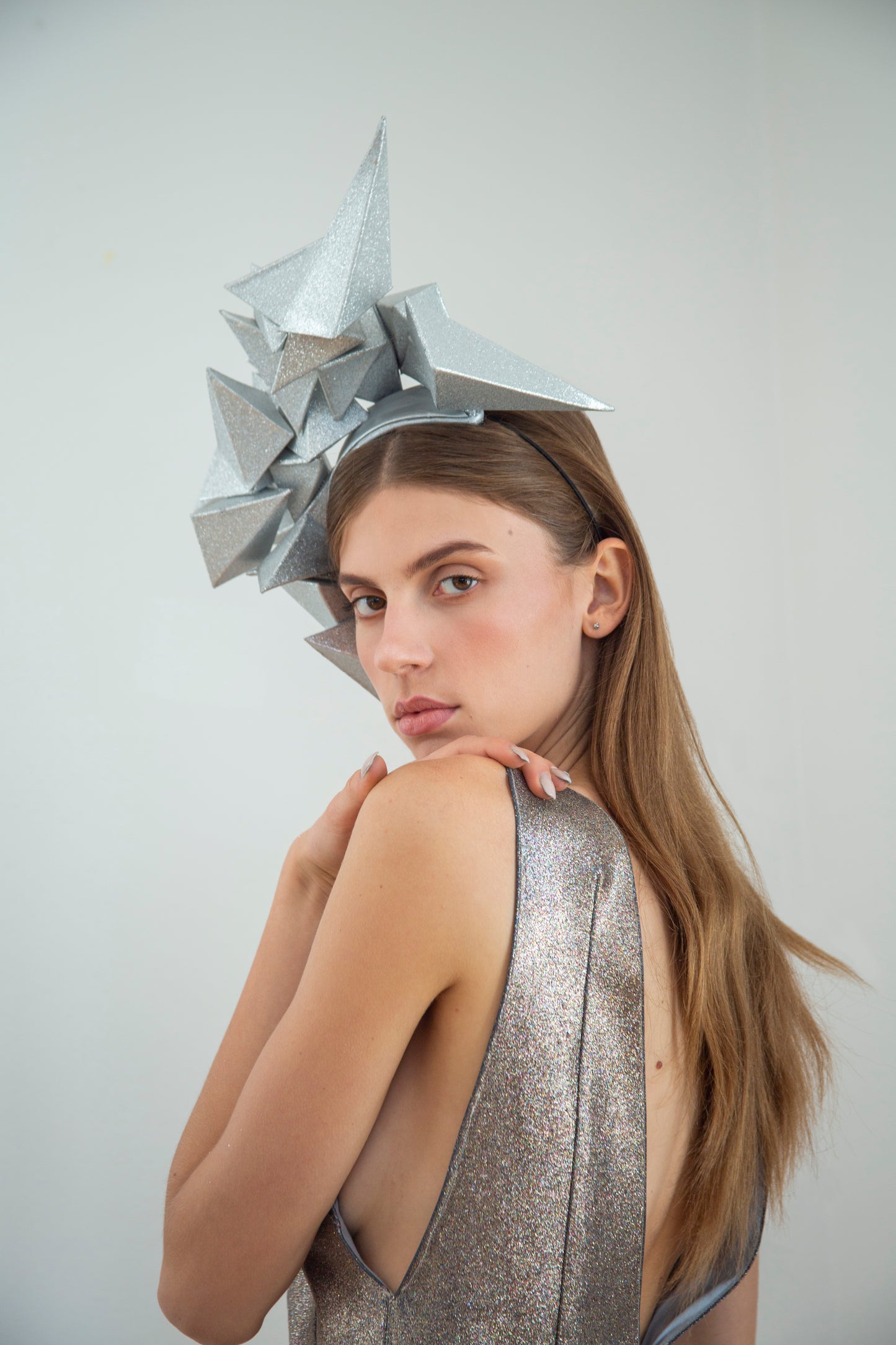 A woman with long brown hair looks over her left shoulder and is wearing a large silver geometric headpiece.