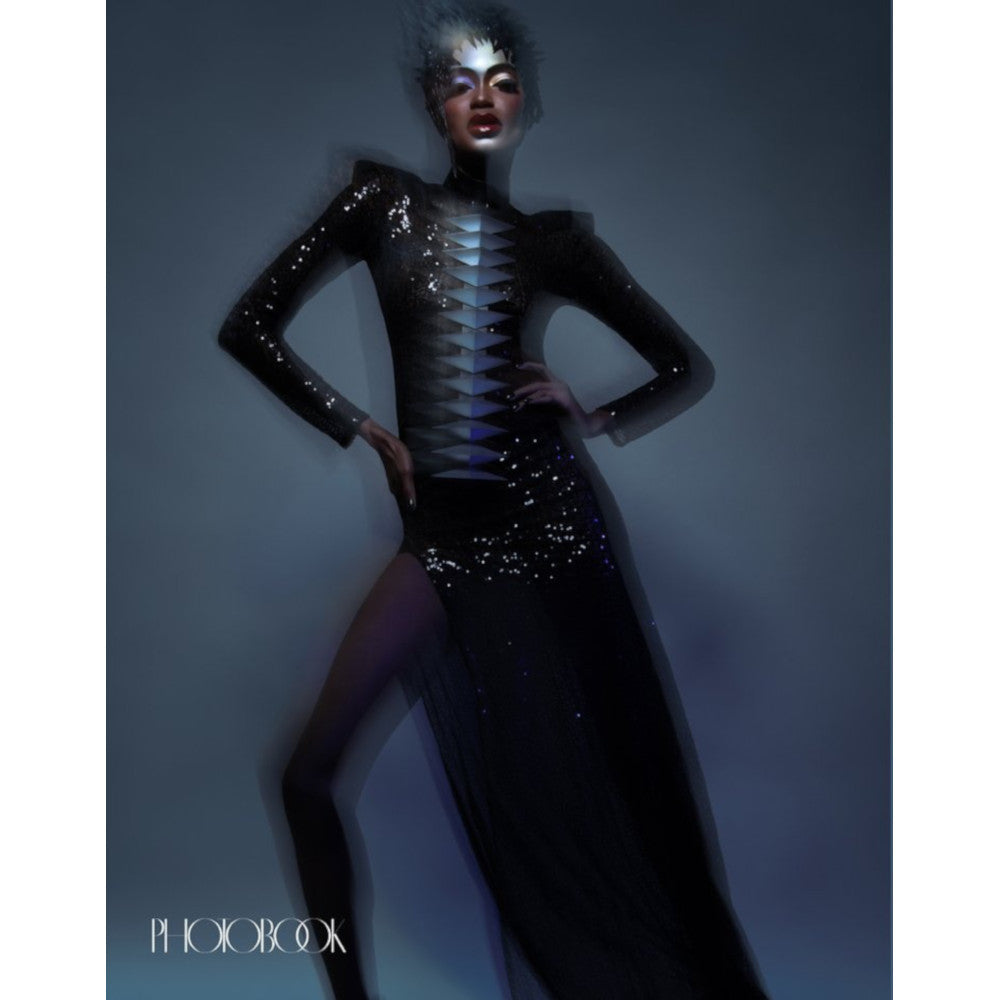 A model poses dramatically in a shimmering black gown with a high slit, adorned with sequins and geometric embellishments.