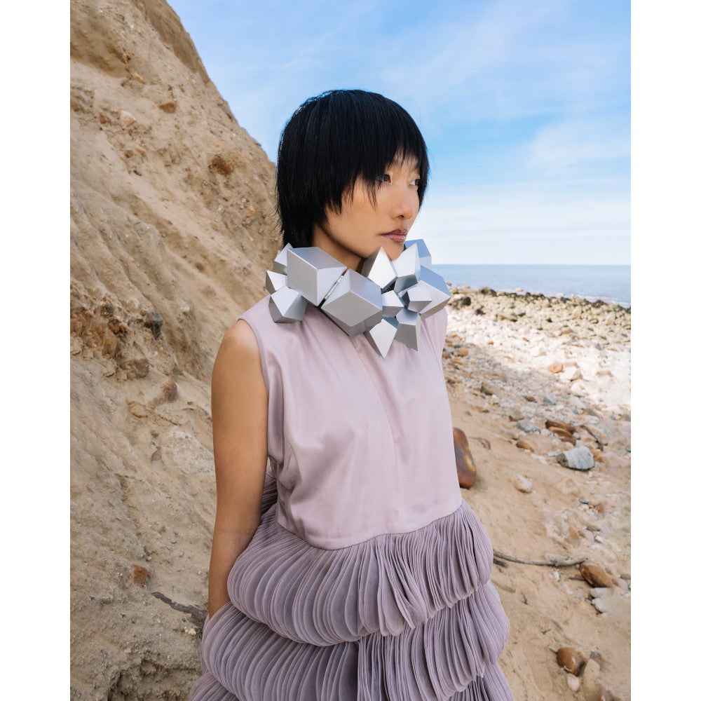 A person wearing a unique geometric silver necklace and a textured lavender dress stands on a sandy beach with a blue sky.