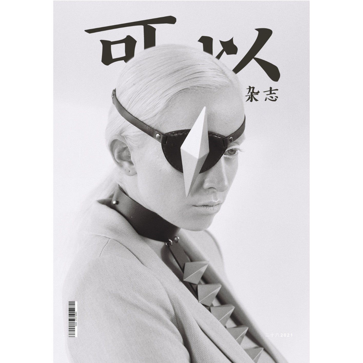 A stylish figure with slicked-back platinum hair wears a black eyepatch with a geometric shape and a tailored gray blazer, featured on a magazine cover.