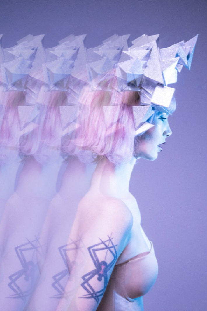 The right side profile of a woman with pink hair standing with a large spider tattoo on her arm wearing a large silver geometric headpiece in front of a purple background.