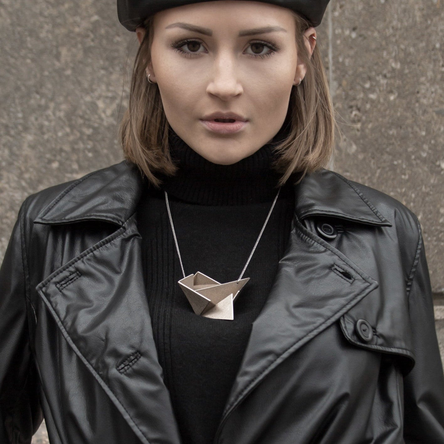 A person wearing a black leather jacket and a black turtleneck, showcasing a unique geometric silver necklace against a textured background.