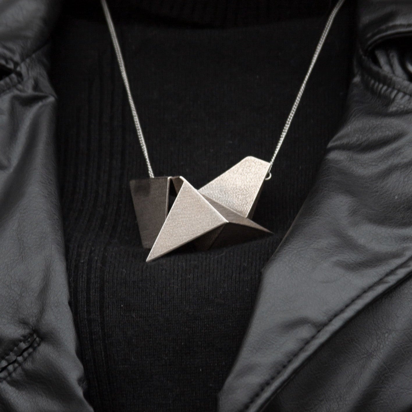 A geometric, origami-inspired pendant in metallic beige hangs from a silver chain, worn over a black turtleneck and leather jacket.