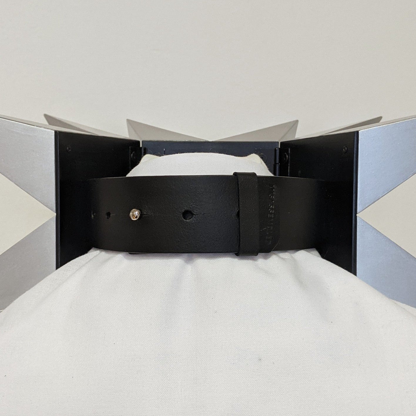 A black leather belt attached to a unique sculptural piece with metallic star-like projections, displayed on a white fabric backdrop.