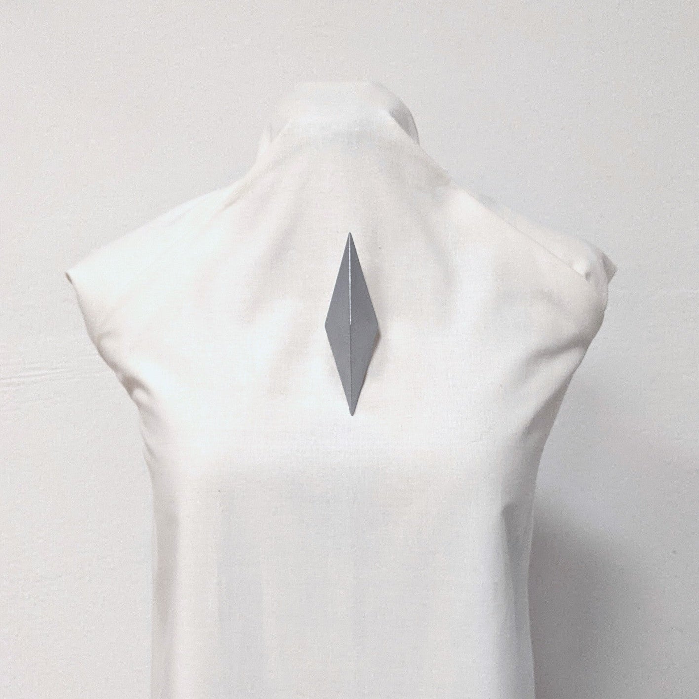 A close-up of a white mannequin displaying a sleek, gray, diamond-shaped brooch pinned to its back.