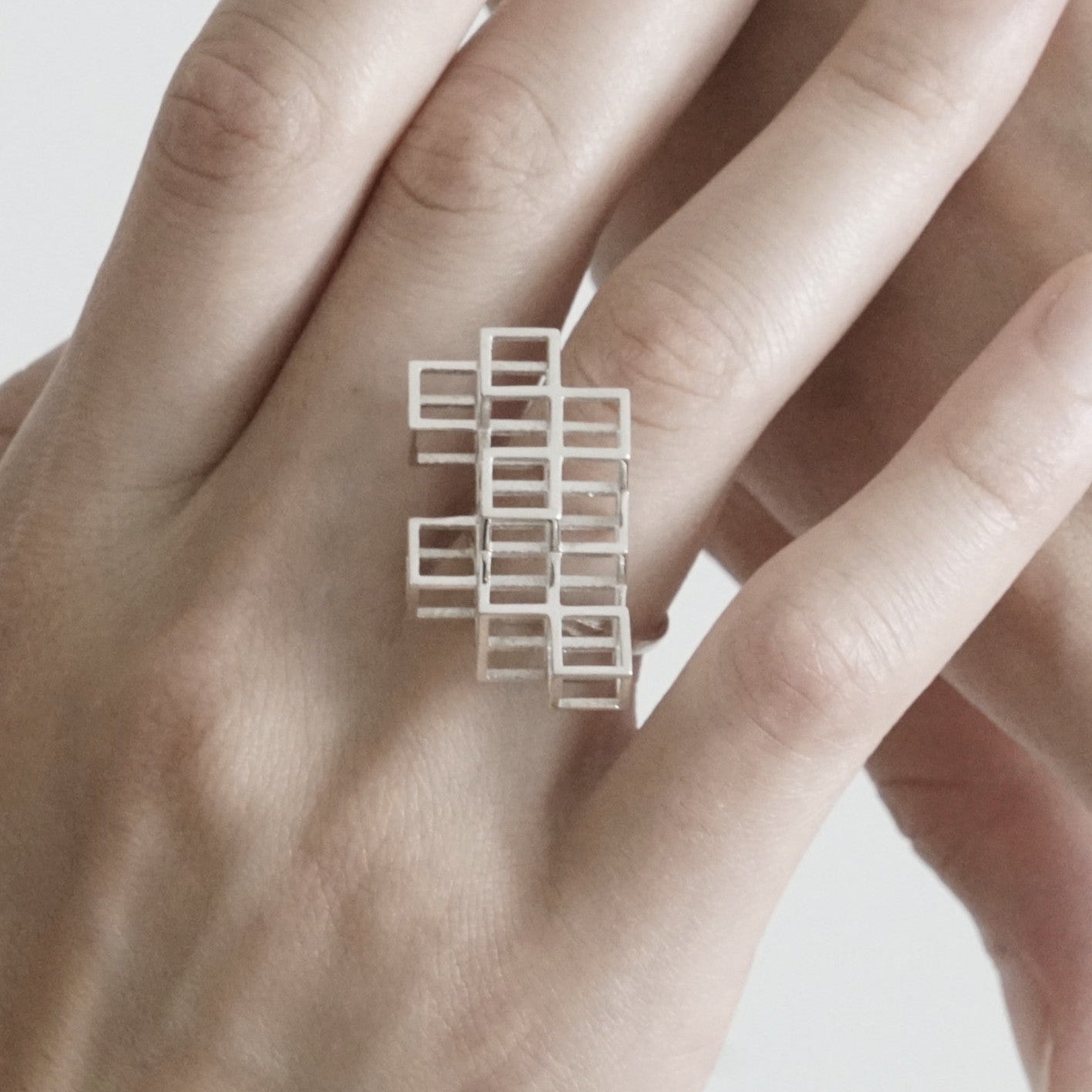 A hand wearing a modern silver geometric ring, featuring overlapping square patterns, against a neutral background.