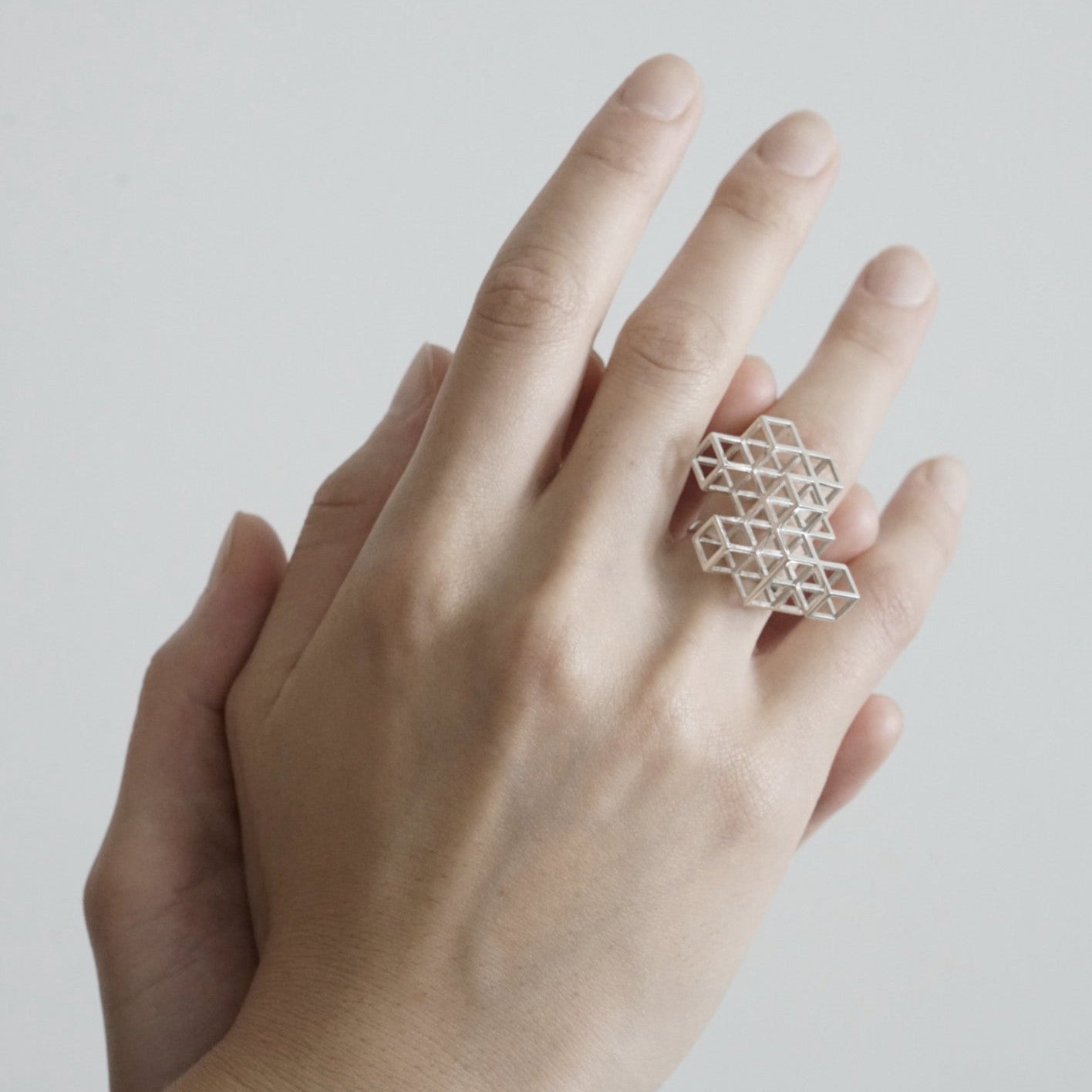 A hand with neatly manicured fingers showcases a geometric silver ring, featuring a lattice of interconnected shapes.