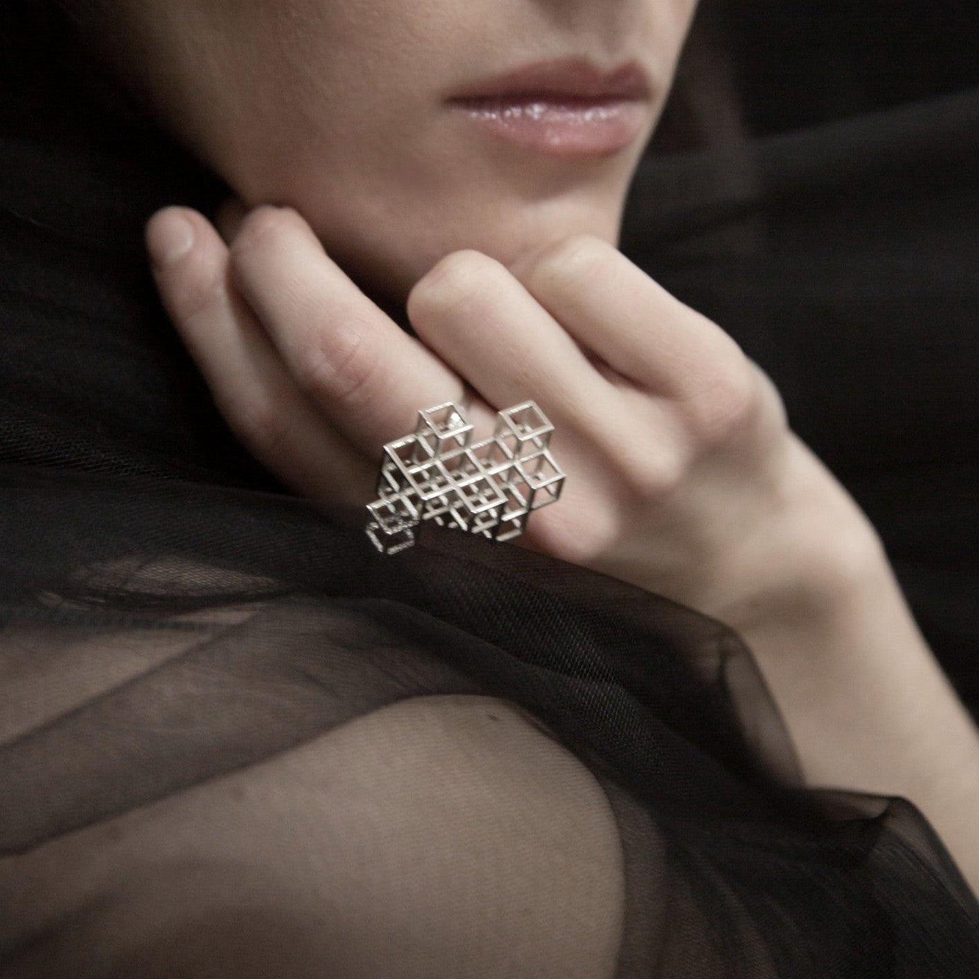 A close-up of a woman's hand with a geometric silver ring, resting against her chin, draped in a sheer black fabric.