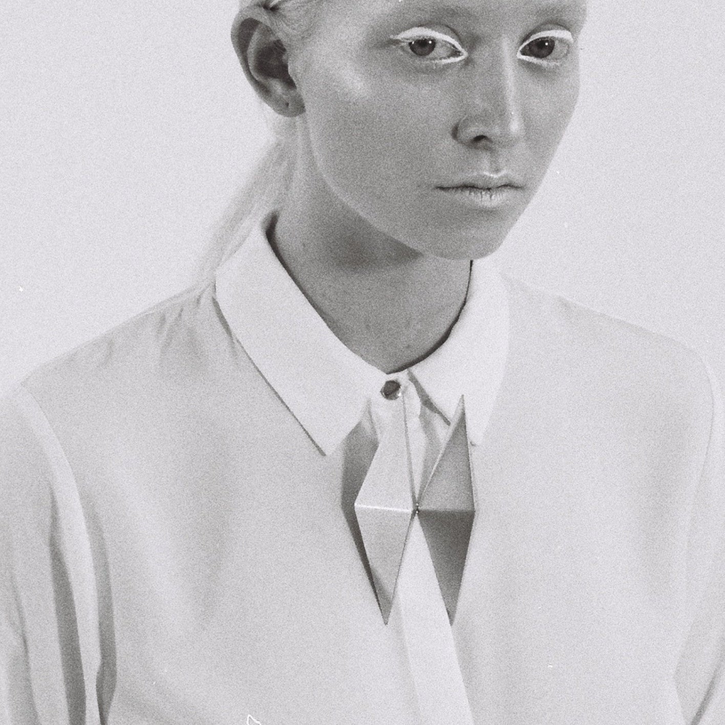 A person wears a crisp white shirt with a geometric silver brooch, set against a simple gray background.