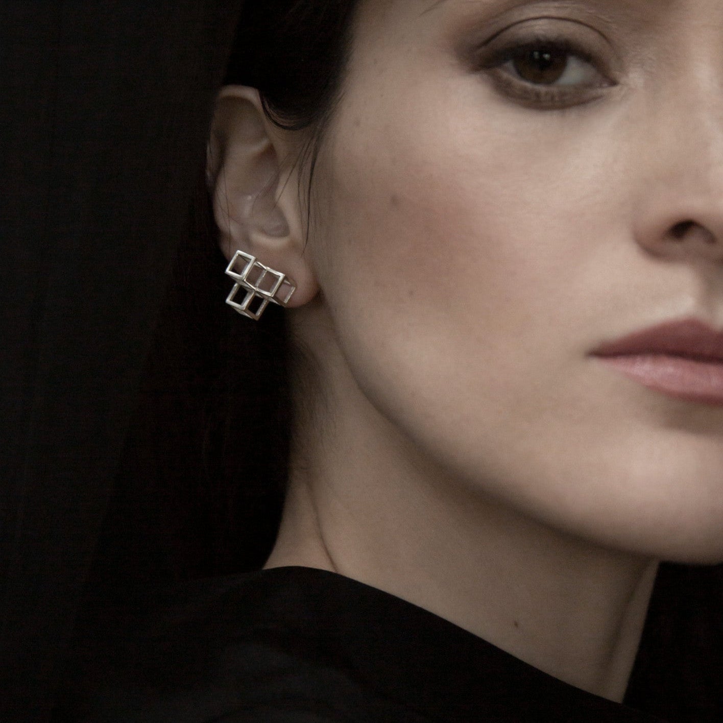 A close-up of a delicate silver geometric earring attached to an ear, against a backdrop of dark fabric.