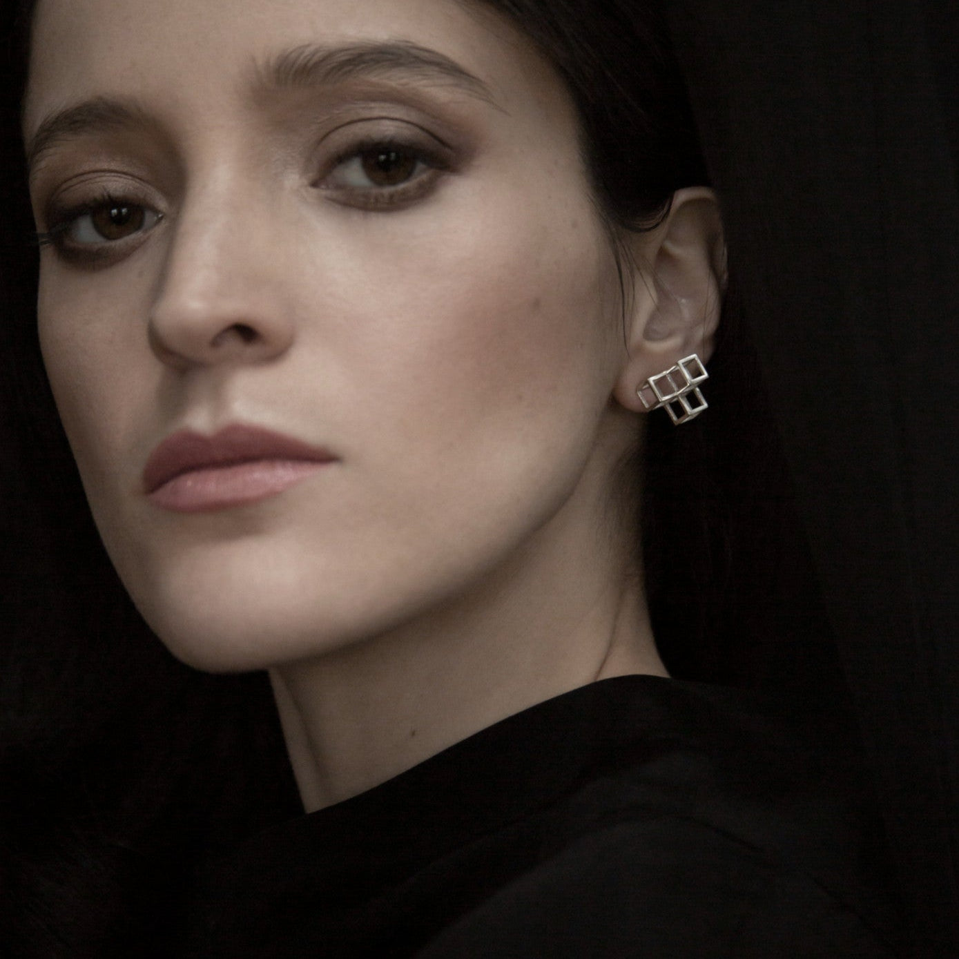 A close-up of an ear adorned with square-shaped silver earrings against a backdrop of dark clothing.
