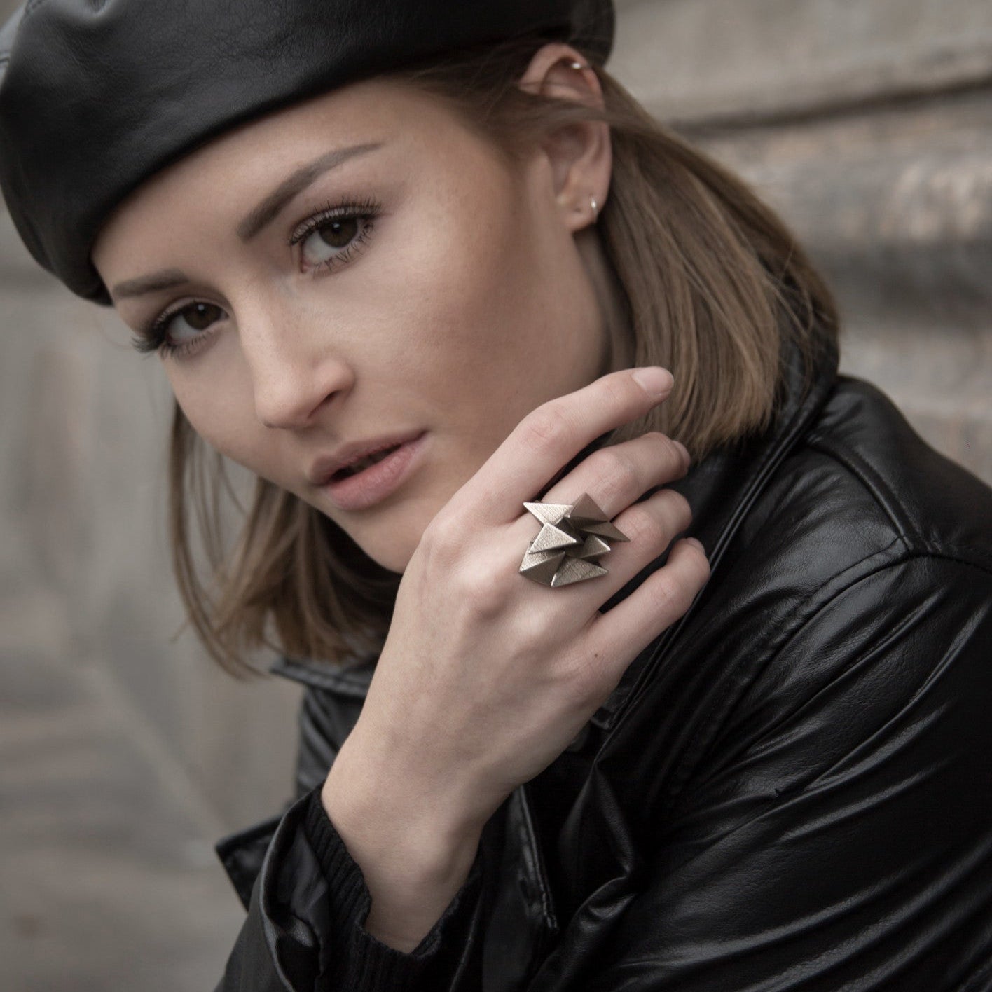 A person wearing a leather jacket and a beret, showcasing a stylish, edgy ring on their hand against a textured background.