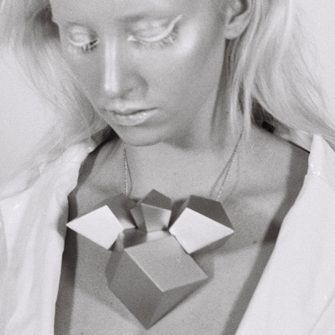 A close-up of an abstract, geometric necklace made of various shapes, worn over a glossy white top against a plain background.