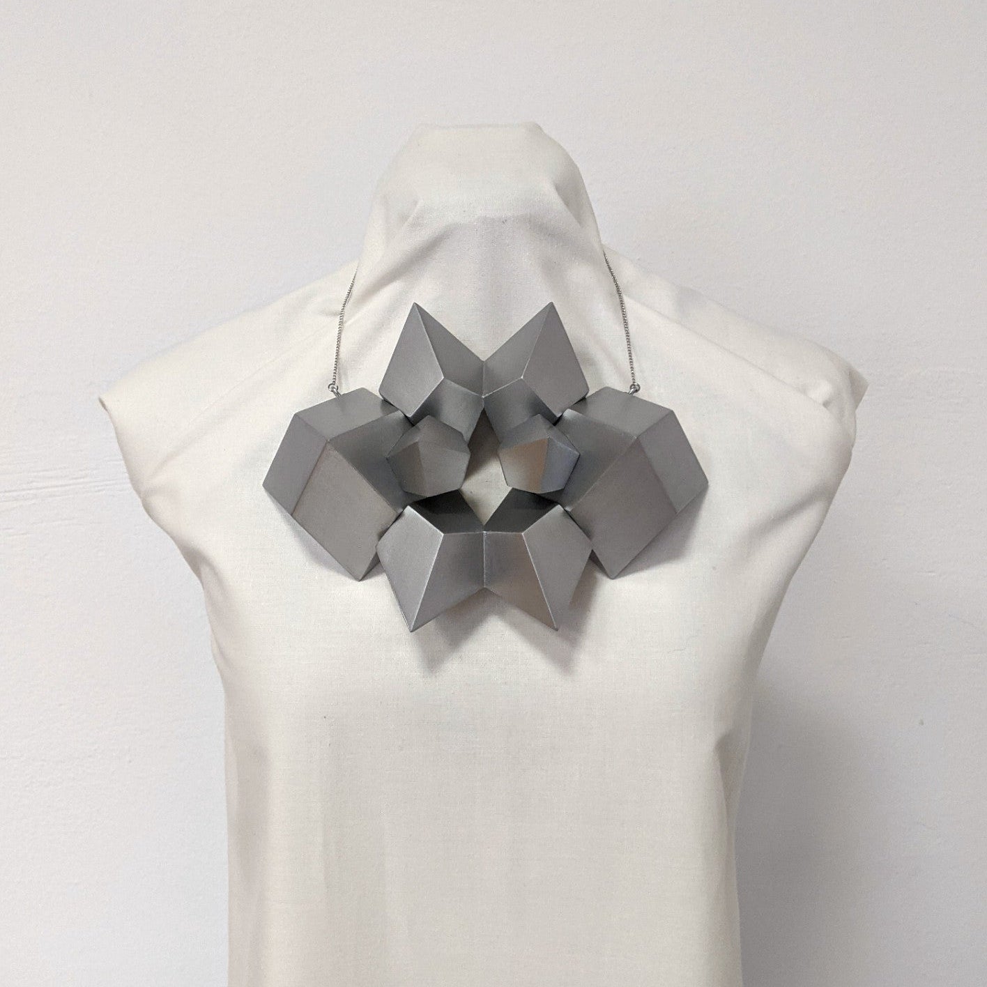 A sculptural silver necklace featuring geometric shapes arranged in an intricate, star-like design on a mannequin bust.