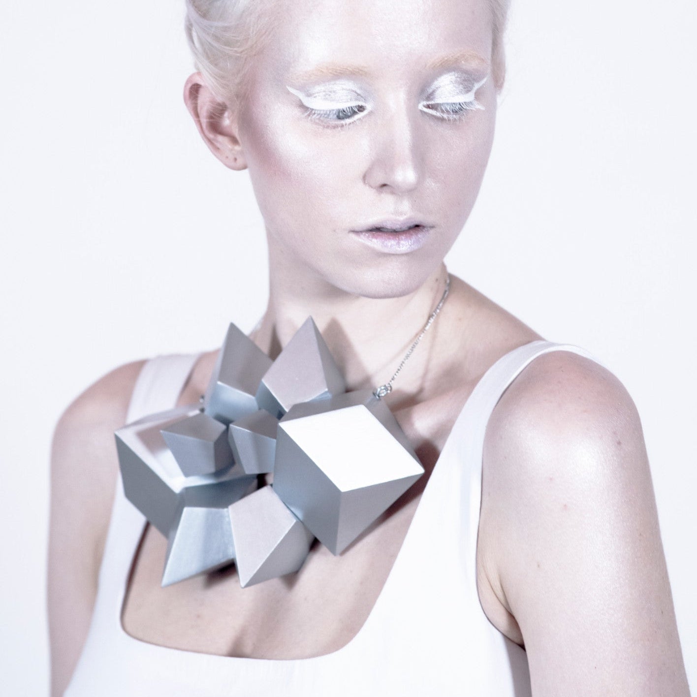 A person wears a large, sculptural silver necklace made of geometric shapes, styled with a simple white top.