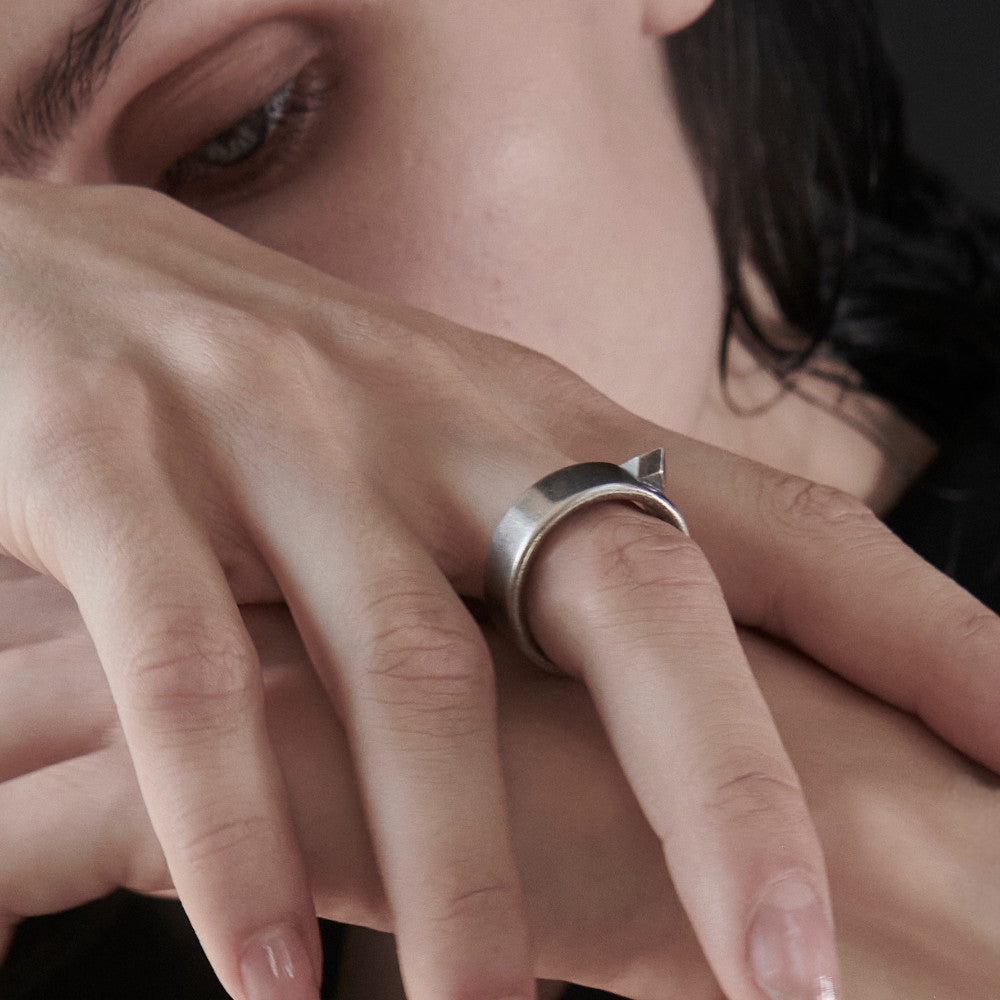 A close-up of two hands intertwining, wearing a silver ring with a geometric shape and showcasing neatly manicured nails against a dark background.