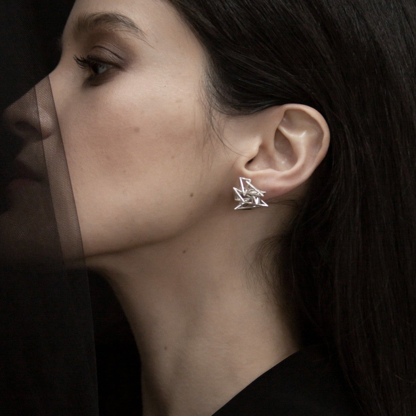 A close-up of a silver geometric earring worn on a woman's ear, complemented by sleek dark hair and a black outfit.