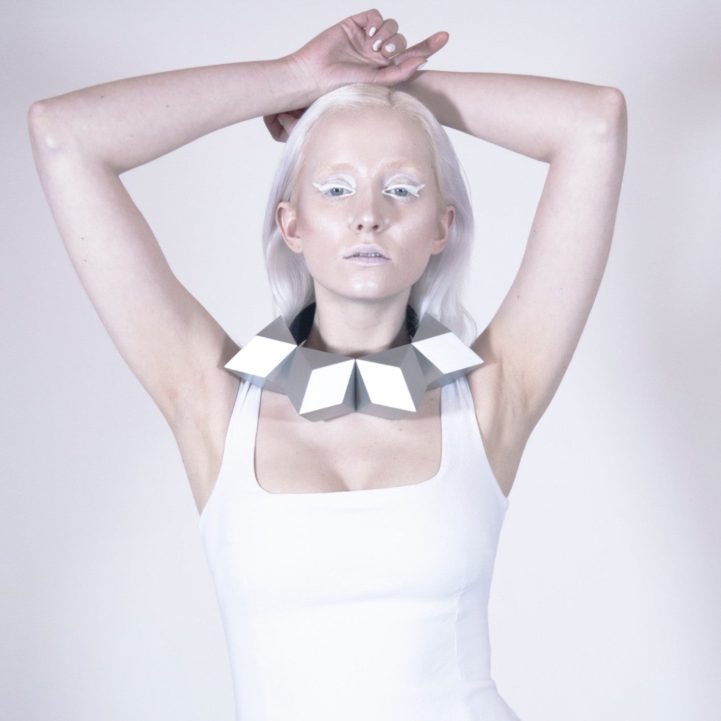 A person with long, platinum hair poses in a white tank top, wearing a striking geometric metallic collar.