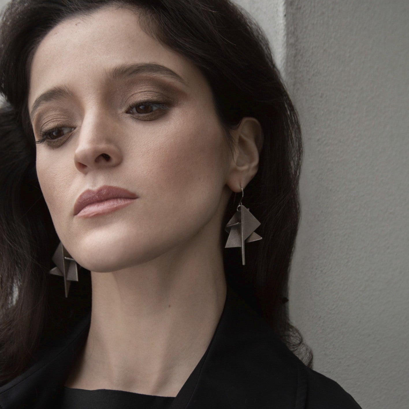 A woman with dark hair standing next to a cement wall wearing distinctive silver geometric earrings, complemented by a black shirt.