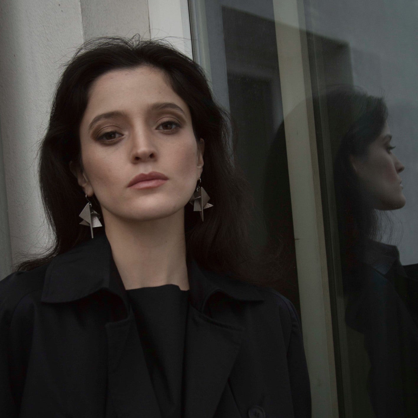 A woman with dark hair standing next to a reflective window wearing distinctive silver geometric earrings, complemented by a black shirt.