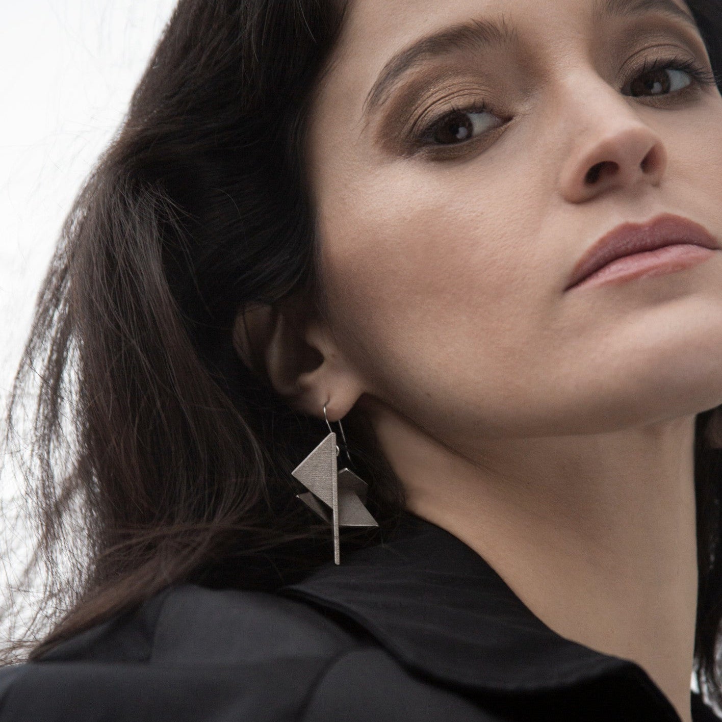 A close-up of a woman with dark hair wearing distinctive silver geometric earrings, complemented by a black shirt.