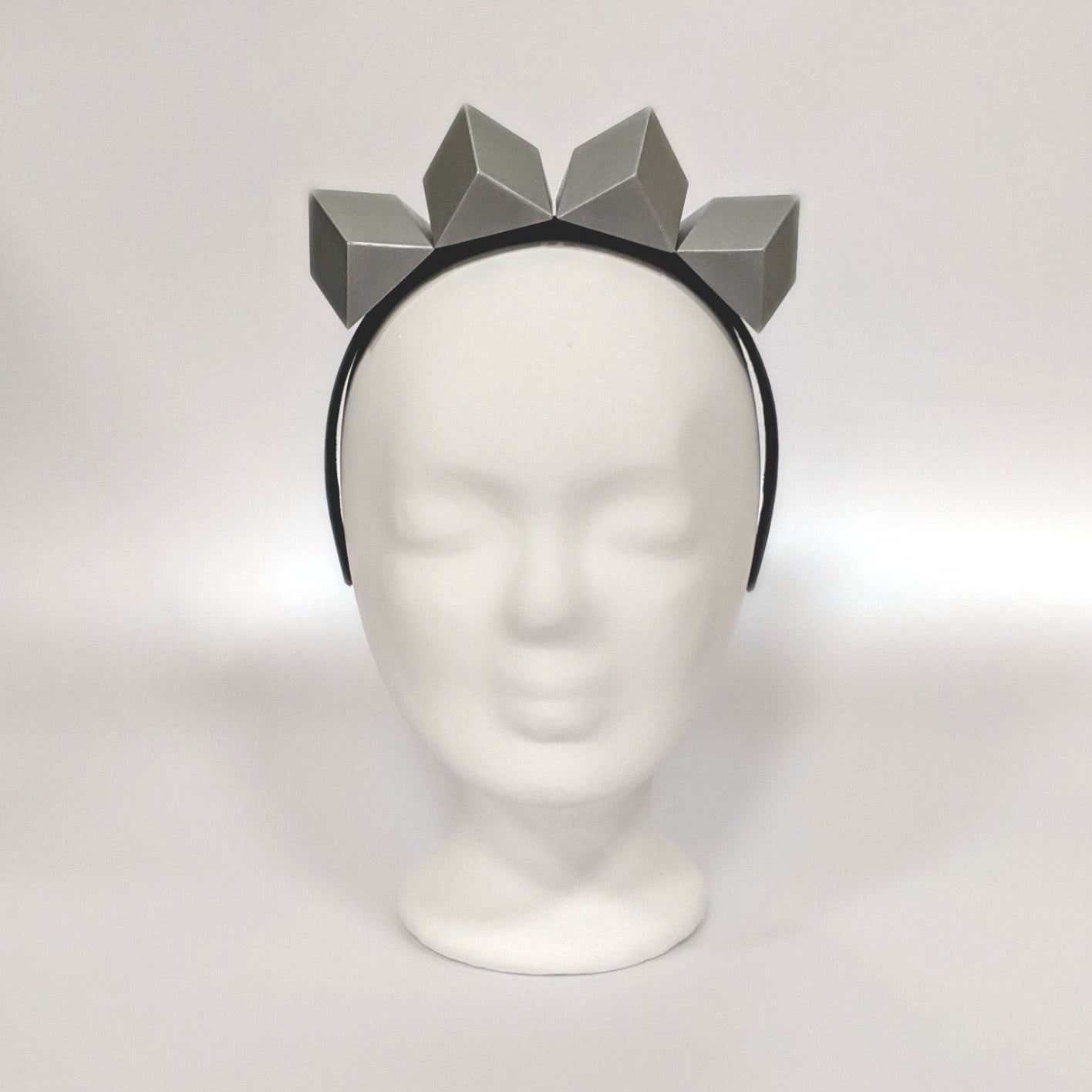 A headband featuring a group of geometric, grey shapes resembling a crown or tiara, poised atop a plain, white display head.