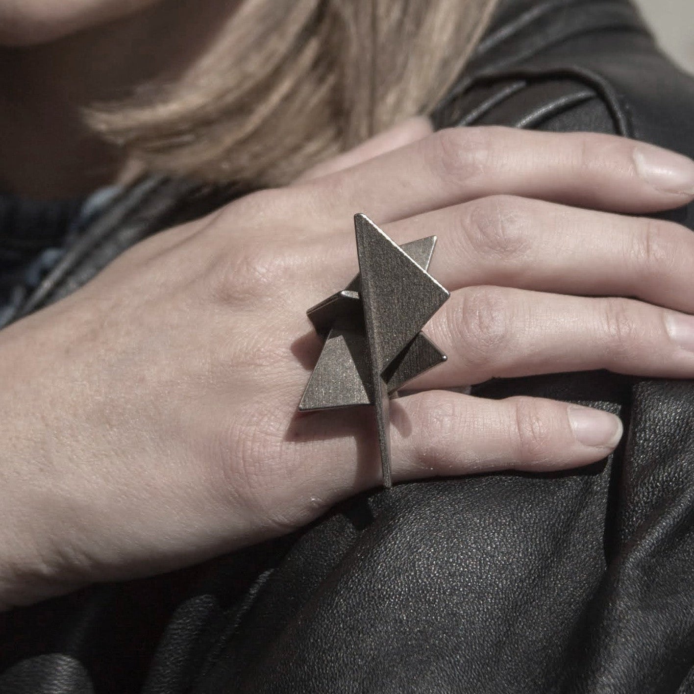 Close-up of a woman's hand resting on her leather jacket, showcasing a unique, geometric ring with metallic surfaces in earthy tones.