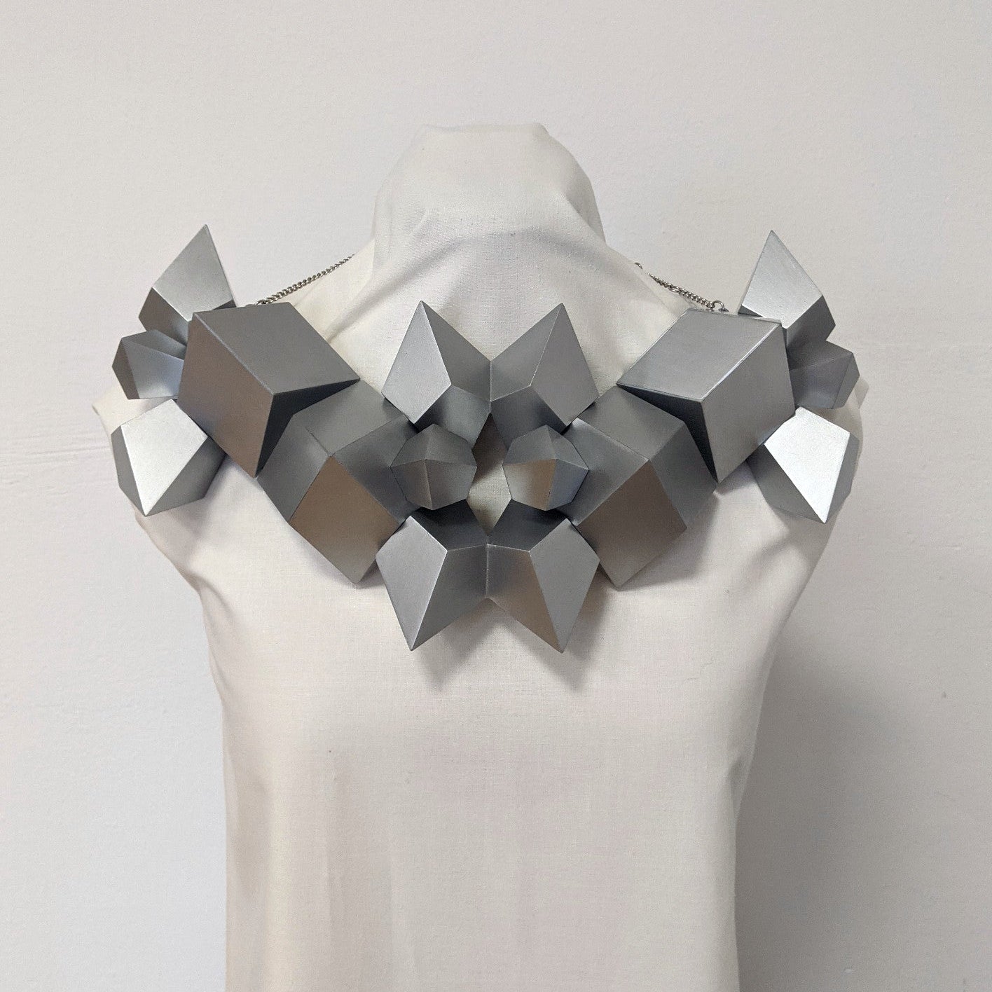 A geometric silver necklace made of angular shapes displayed on a mannequin against a plain background.