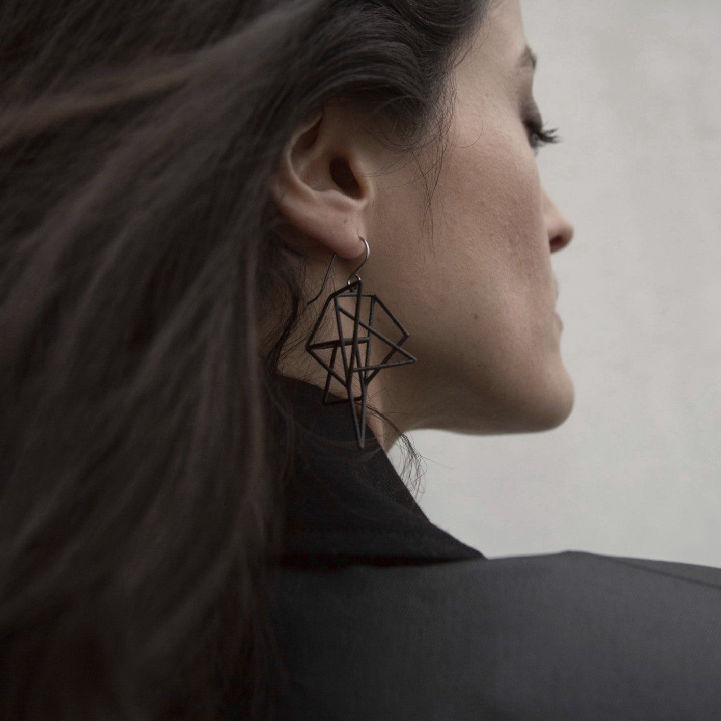 A woman with long dark hair wears bold geometric earrings, styled in a chic black jacket, against a minimalist background.
