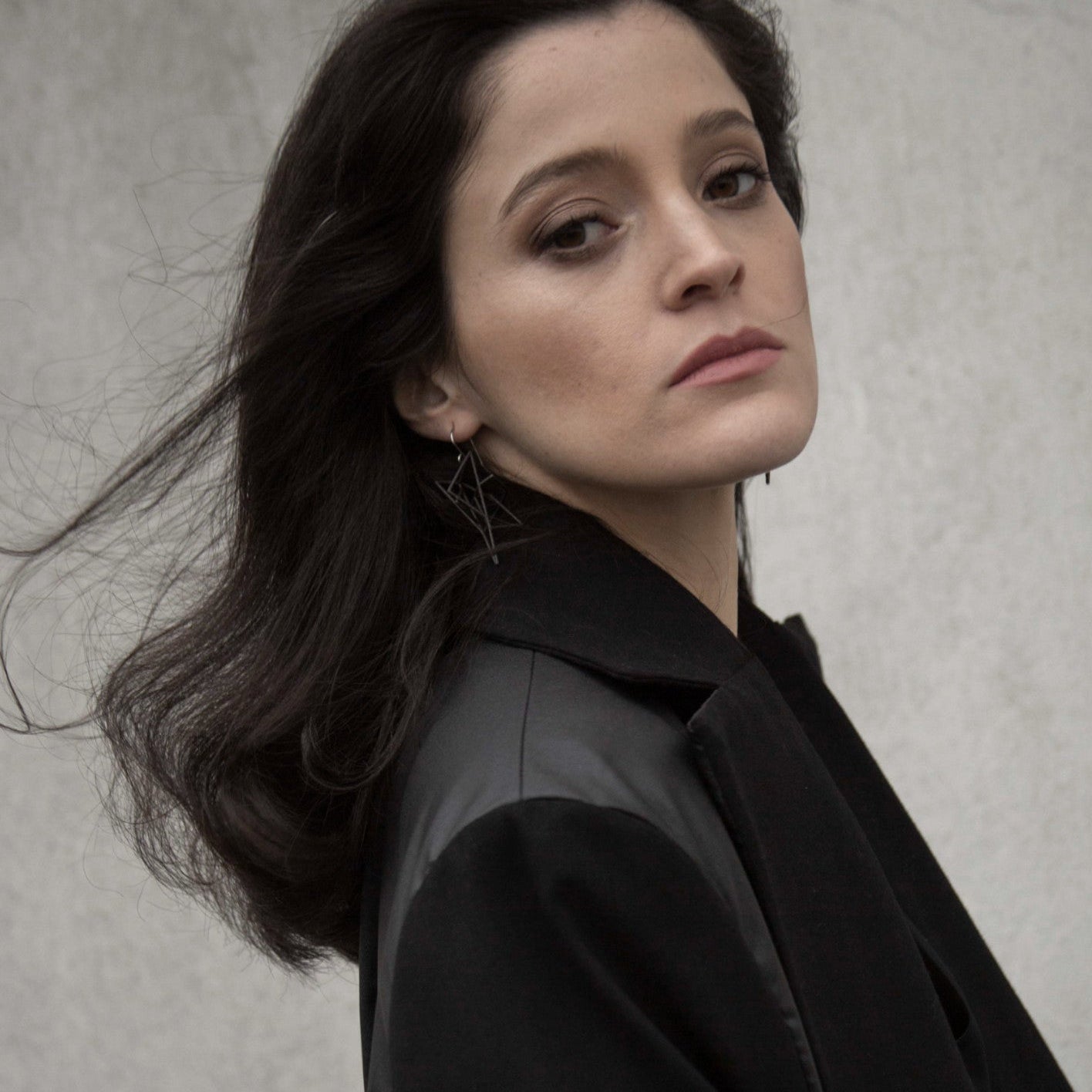 A woman with long dark hair wears bold geometric earrings, styled in a chic black jacket, against a minimalist background.