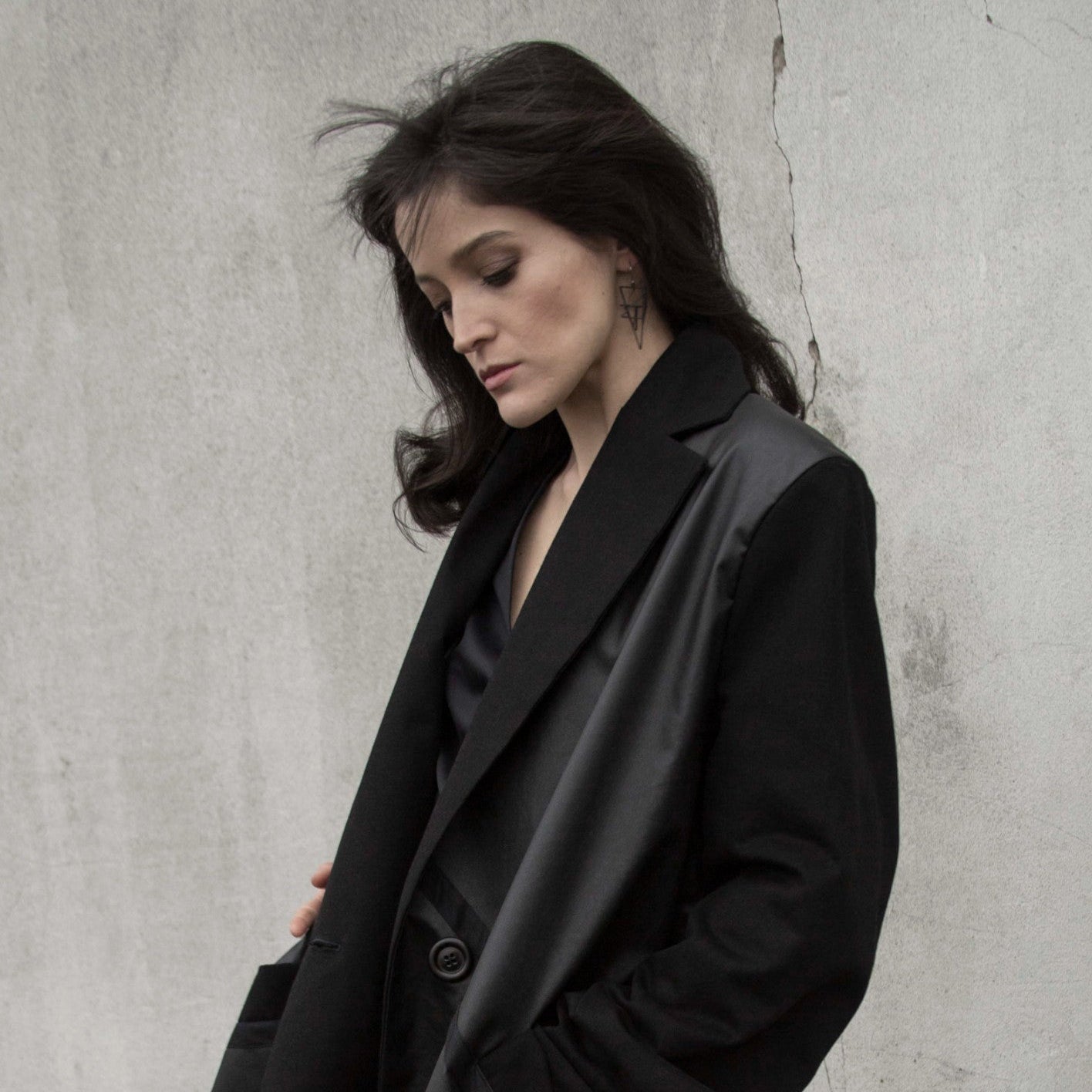 A woman with long dark hair wears bold geometric earrings, styled in a chic black jacket, against a minimalist background.