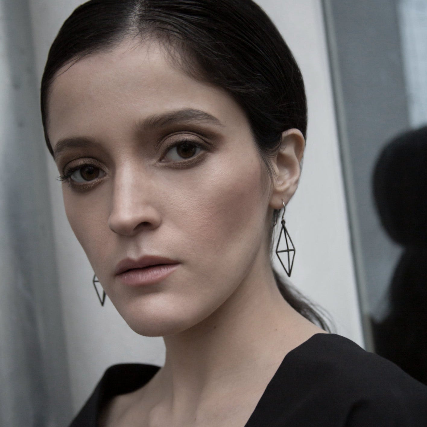 A close-up of a person wearing a stylish black outfit and geometric earrings, set against a neutral background.