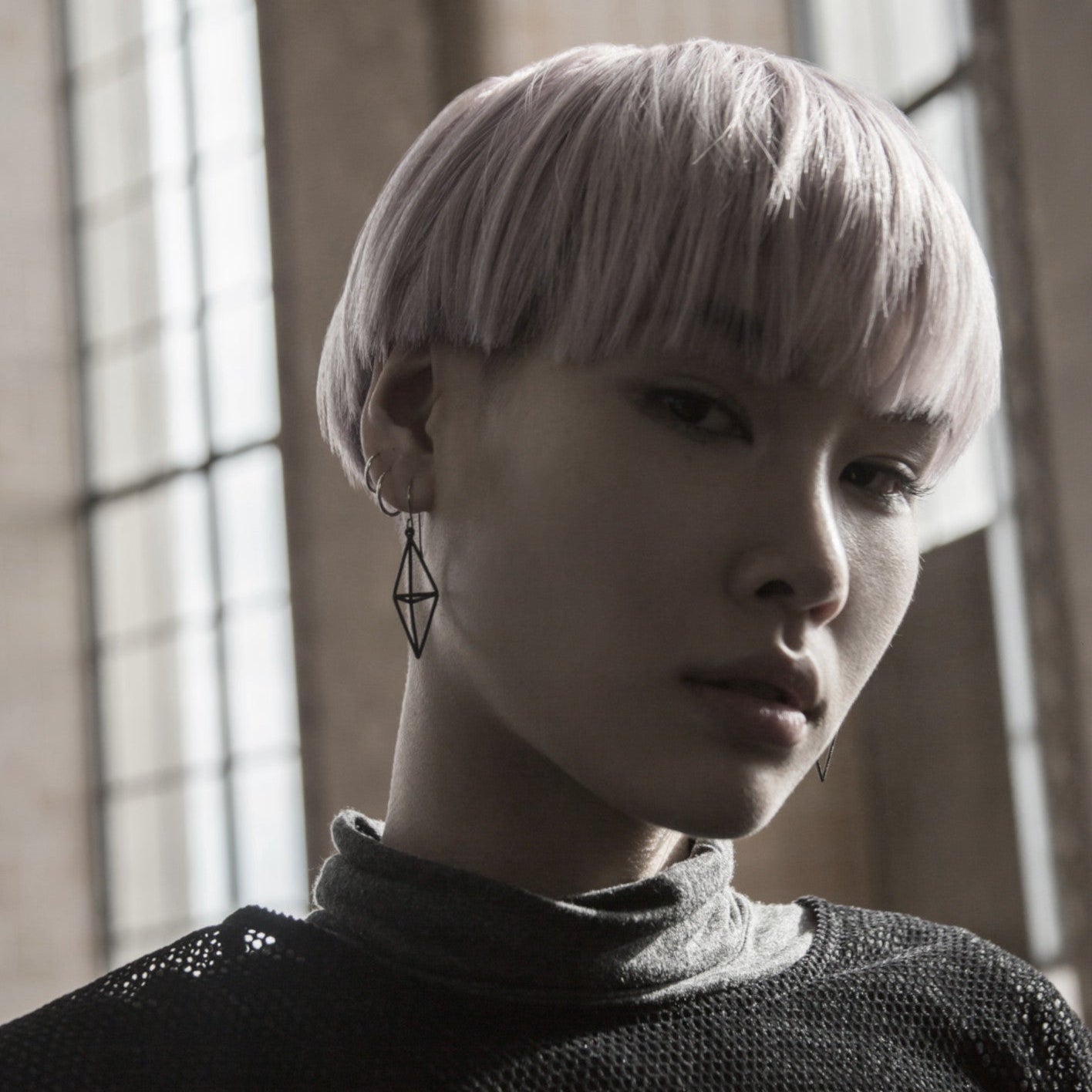 A person with short, pastel pink hair wears a geometric earring and a gray turtleneck top, captured in soft natural lighting.