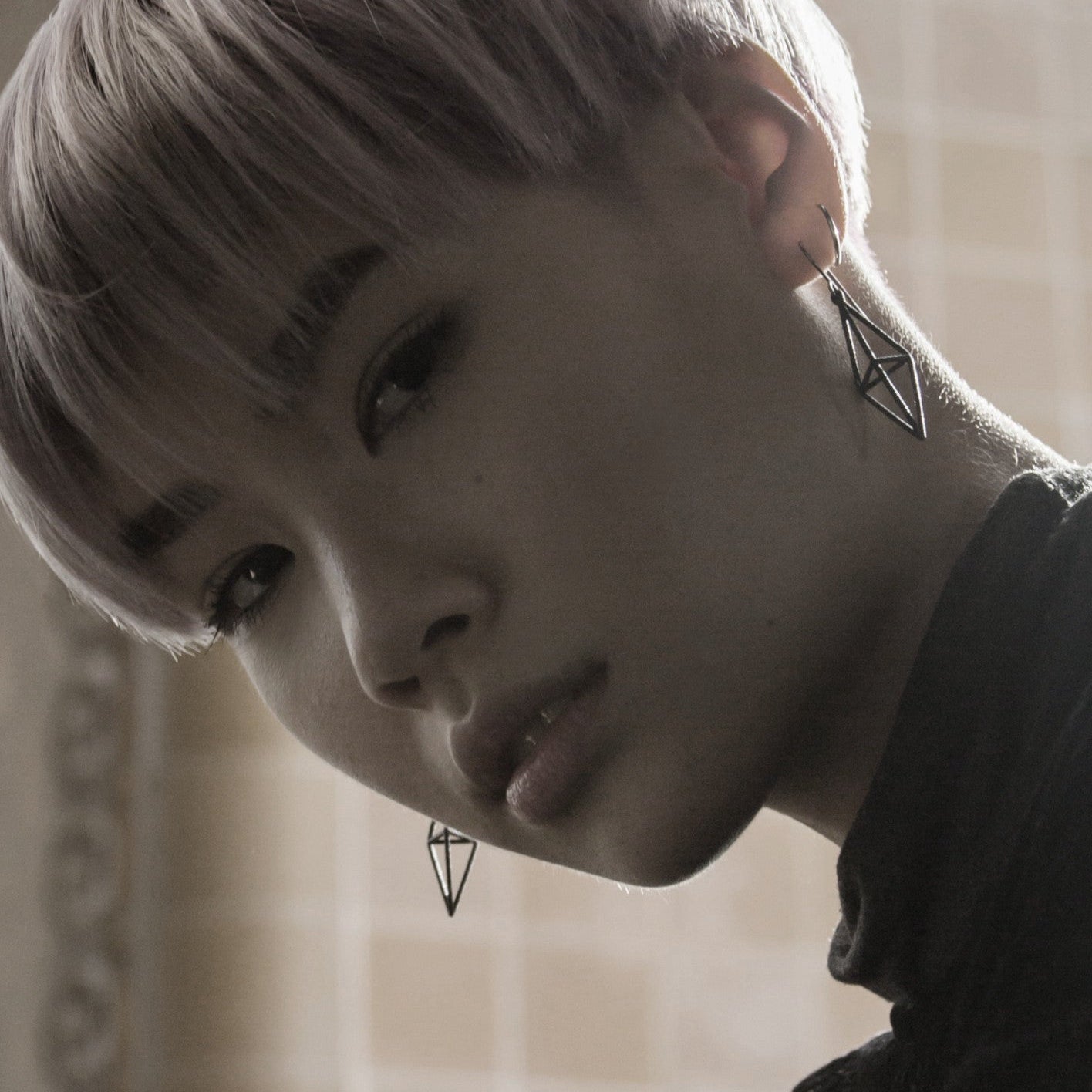 A close-up of a person with pink hair, wearing geometric earrings and a black turtleneck, softly lit against a tiled background.
