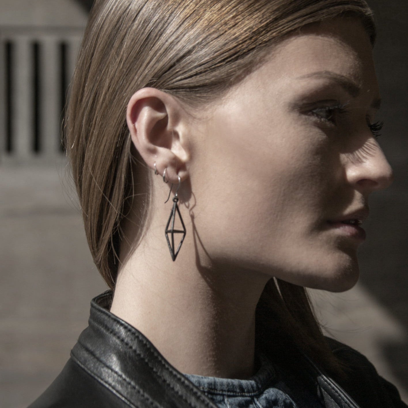 A close-up of a person's ear showcasing stylish geometric earrings and smooth, straight golden hair, with a shadow obscuring the face.
