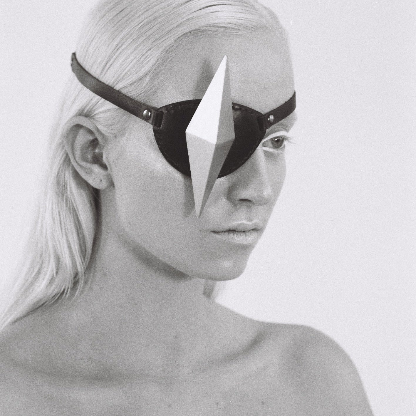 A close-up of a person with long, straight, platinum blond hair, wearing a black eyepatch with a silver geometric shape and showcasing a bare shoulder in black and white.